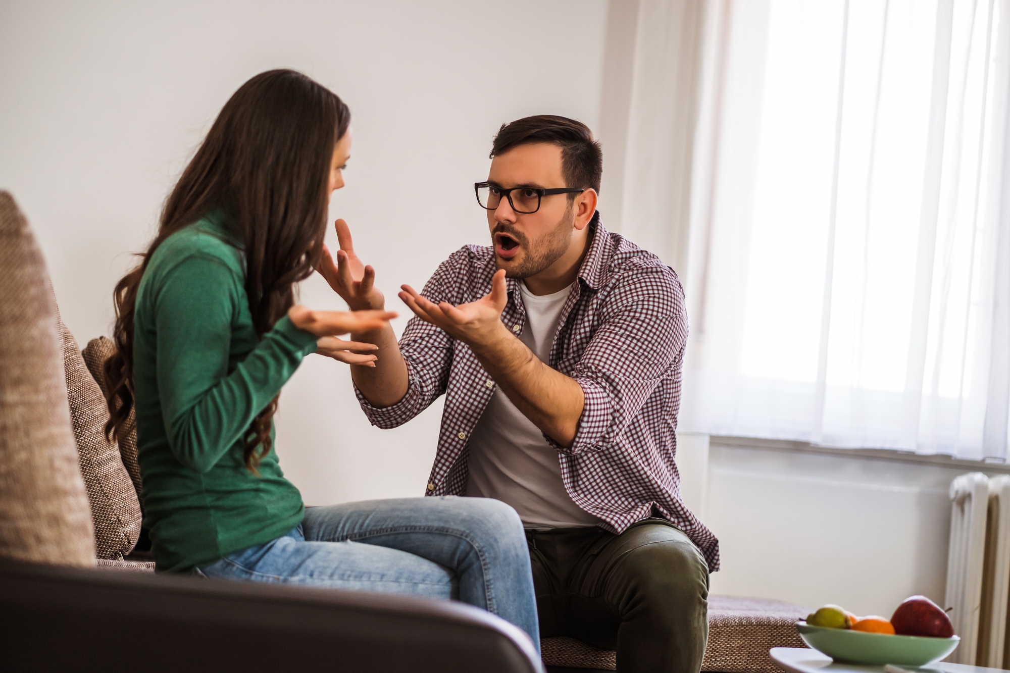 A man and woman are sitting on a couch in a heated discussion. The man, wearing glasses and a checkered shirt, gestures passionately with his hands and has an angry expression. The woman, in a green sweater and jeans, gestures back, looking distressed.