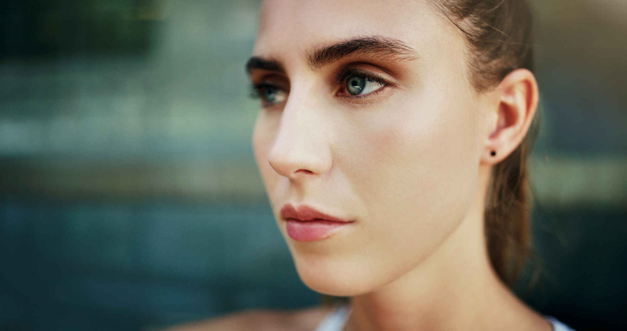 Close-up of a person with long hair pulled back, looking to the right with a neutral expression. The background is blurred, drawing focus to the person's face which shows clear, smooth skin and subtle makeup.