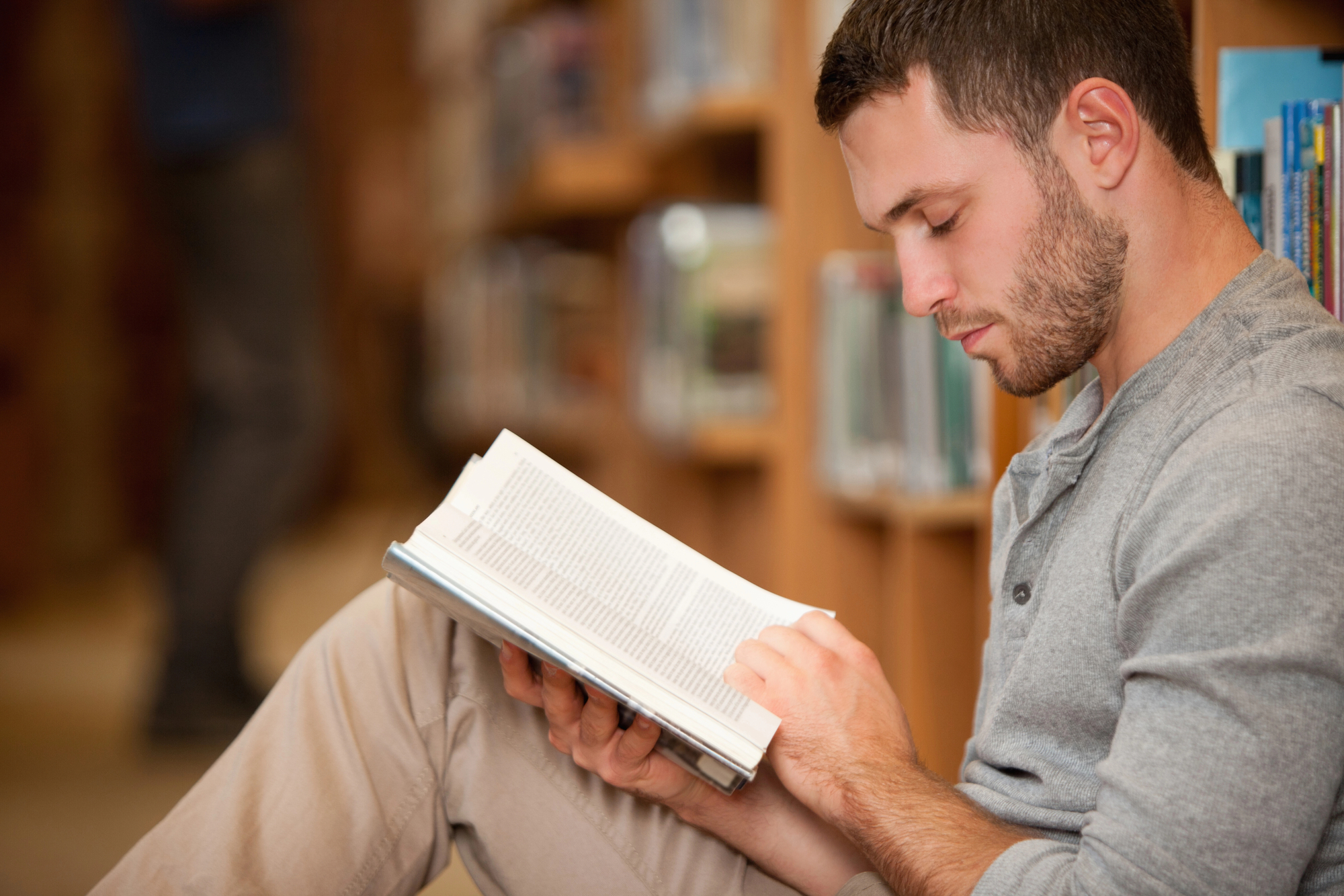 A man with short hair and a beard is sitting on the floor in a library, reading a book. He is dressed casually in a gray long-sleeve shirt and beige pants. Shelves filled with books are visible in the background.