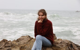 A woman with curly hair wearing a red sweater and blue jeans sits on a rocky shoreline. She rests her chin on her hand and looks at the camera with a slight smile. The ocean waves crash behind her on a cloudy day.