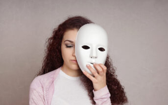 A young woman with long, curly hair holds a white, featureless mask in front of her face, slightly off to one side, revealing her closed eyes. She is wearing a light pink cardigan over a white shirt and stands against a neutral gray background.