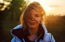 A woman with light brown hair, partially covering her face, stands outdoors at sunset. She is wearing a white jacket with a black inner lining. The background is blurred, featuring green foliage and a glowing sun near the horizon.