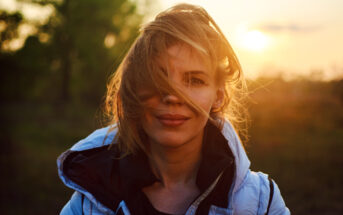 A woman with light brown hair, partially covering her face, stands outdoors at sunset. She is wearing a white jacket with a black inner lining. The background is blurred, featuring green foliage and a glowing sun near the horizon.