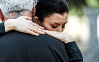 A woman with dark hair embraces an older man with gray hair. Her eyes are closed, and she rests her head on his shoulder. Their hands are visible, and they appear to be sharing a tender moment. The background is blurred, highlighting the emotional connection.