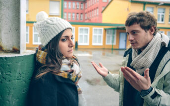 Two people are outside, engaged in a serious conversation. One person, wearing a white beanie and a scarf, leans against a green pillar and looks away. The other person, wearing a scarf and gesturing with both hands, speaks animatedly. They are in a schoolyard area.