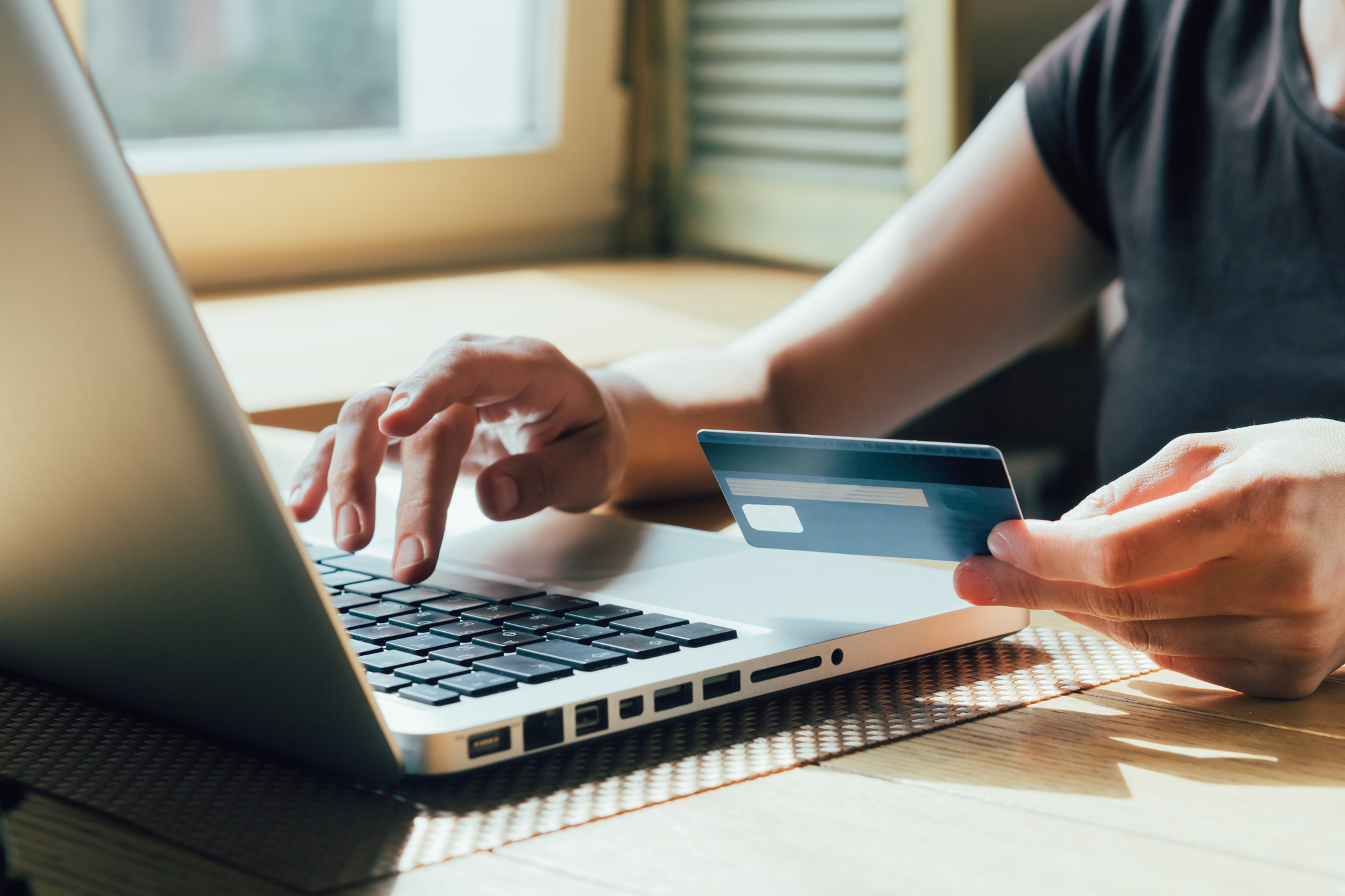 A person sits at a table using a laptop while holding a credit card in their left hand. Sunlight streams through a window in the background, illuminating the workspace. The scene suggests online shopping or electronic payment.