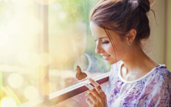 A woman with brown hair in a bun smiles while looking at a butterfly resting on a window. She is indoors, and sunlight filters through the window, casting a warm glow. She is wearing a floral print blouse and gently touching the glass near the butterfly.