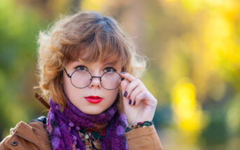 A person with wavy, light brown hair is wearing round glasses, a purple scarf, and a brown coat. They are tilting their head slightly and looking forward, holding the glasses with one hand. The background is blurred with autumn colors.