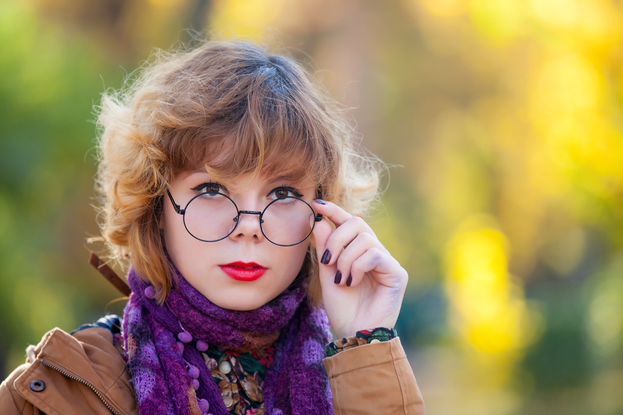 A person with wavy, light brown hair is wearing round glasses, a purple scarf, and a brown coat. They are tilting their head slightly and looking forward, holding the glasses with one hand. The background is blurred with autumn colors.