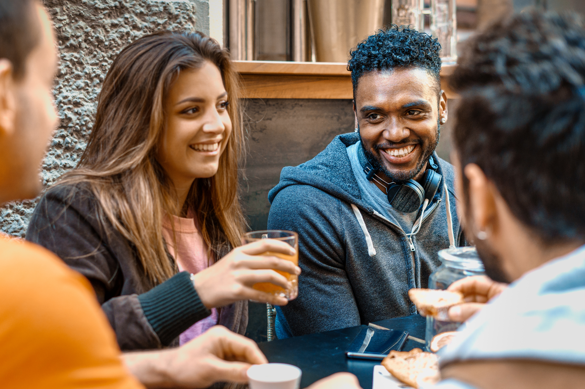 A group of four friends is sitting at an outdoor café table, enjoying drinks and food. They are engaged in conversation and laughing. One person holds a glass of orange juice, while another has a cup of coffee. There are plates of food on the table.