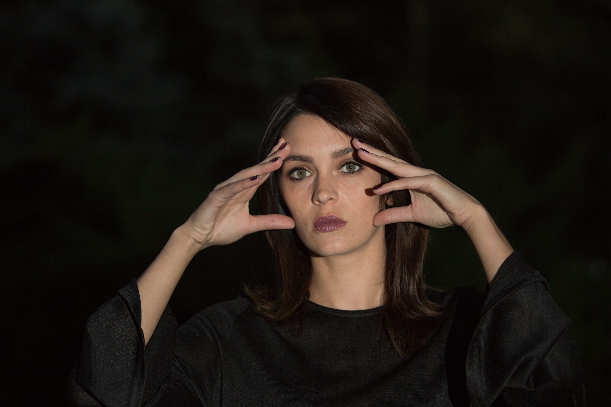 A woman with shoulder-length brown hair and green eyes is posing against a dark, blurred background. She is wearing a black long-sleeve top and holding her hands near her face, with fingers spread and a serious expression.