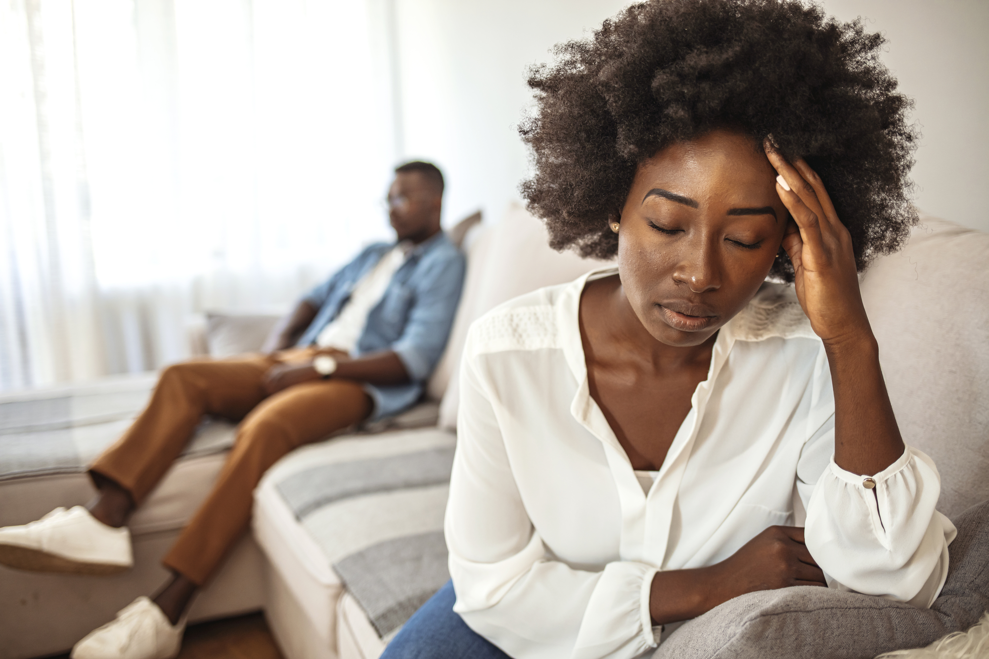 A woman with an afro hairstyle sits on a couch, resting her head on her hand in apparent distress or deep thought. In the background, a man sits on another part of the couch, partially blurred, looking away from the woman. The room has soft, natural lighting.