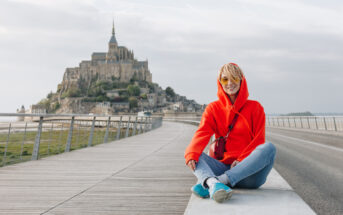 A person in a red hoodie and blue shoes sits on a wooden pathway with Mont Saint-Michel in the background. The sky is slightly overcast, and the pathway leads directly to the historic island commune. The person appears to be smiling and enjoying the view.