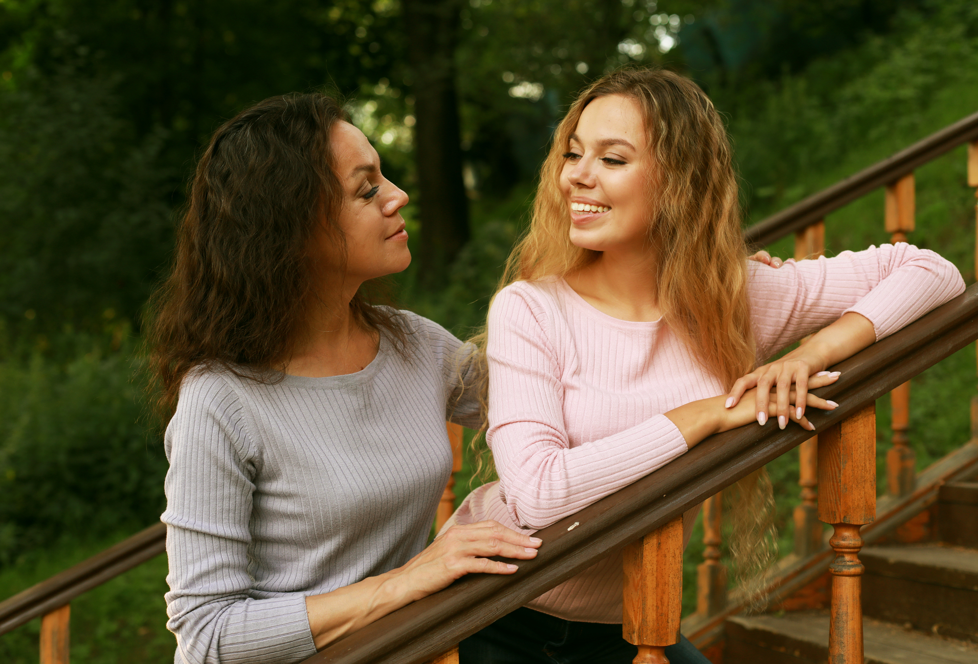 Two women stand on outdoor wooden stairs, surrounded by lush greenery. They smile at each other warmly; one has curly dark hair and wears a light purple sweater, while the other has wavy blonde hair and is dressed in a pink sweater.