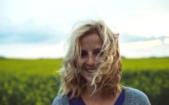 A person with shoulder-length blonde hair smiles while standing in a field of green grass and yellow flowers. The sky is partly cloudy, and the wind is blowing the person's hair across their face.