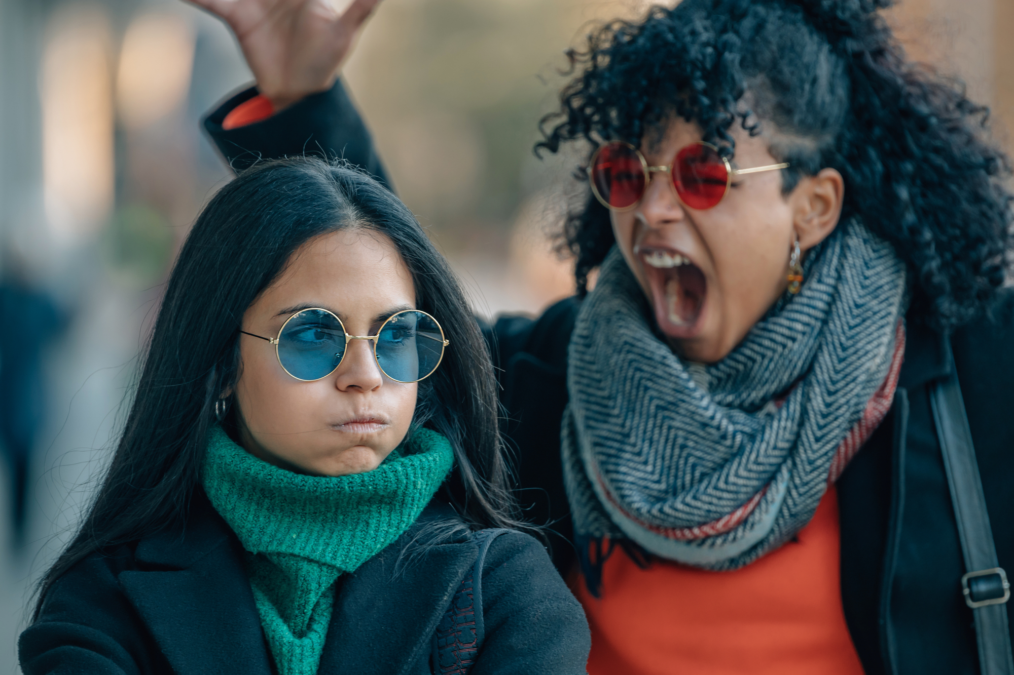 Two women are outdoors. The woman in the foreground is wearing blue sunglasses, a green turtleneck, and a serious expression. The woman in the background has curly hair, red sunglasses, and an exaggerated yelling expression, with one hand raised.