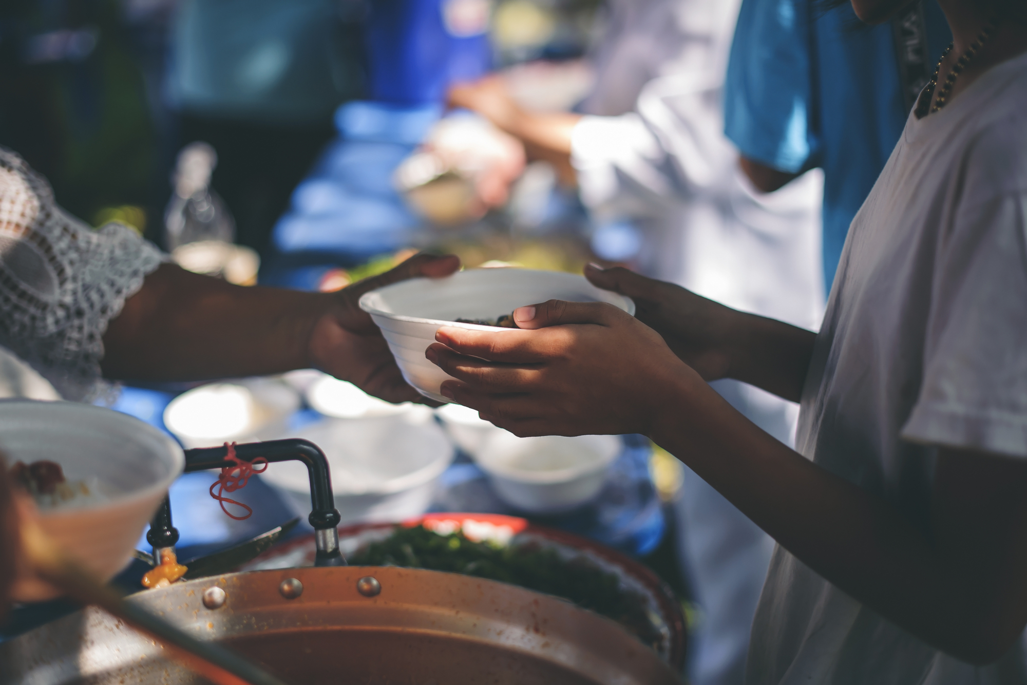 A person hands a white bowl of food to another person at an outdoor event. The scene is bustling, with more people in the background. The setting appears to be a communal meal or food distribution, with various dishes and utensils visible on a table.