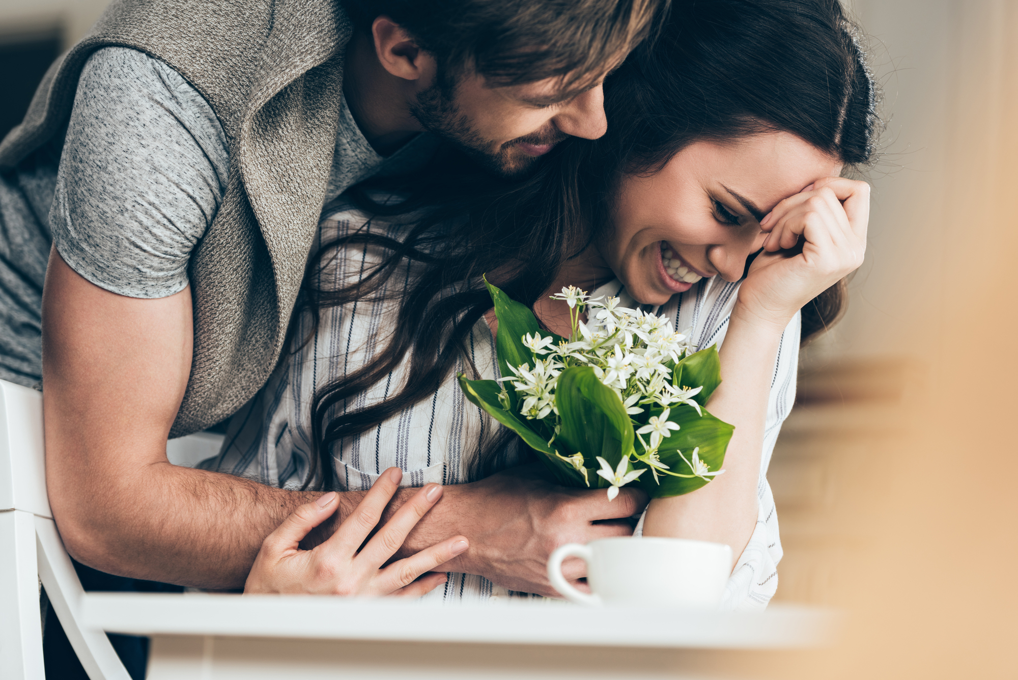 A man embraces a smiling woman from behind as she holds a bouquet of flowers. They both appear happy and affectionate. The woman, partially hiding her face with her hand, has long dark hair and is wearing a striped shirt. A coffee cup is seen on the table in front of them.