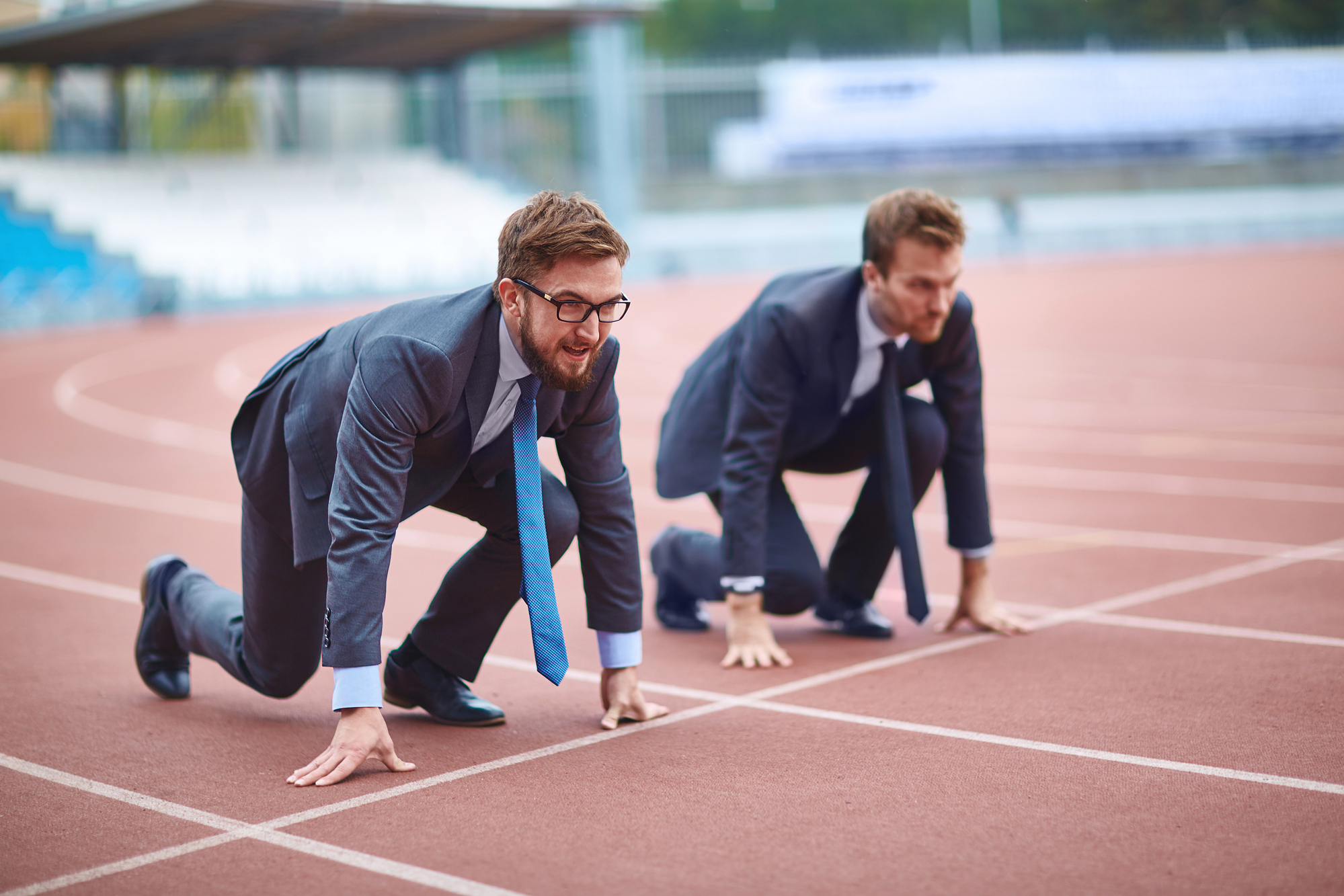 Two men dressed in business suits and ties are crouched at the starting line on a running track, poised to race. The scene blends professional attire with athletic competition as they prepare for a sprint. The background features empty stadium bleachers.