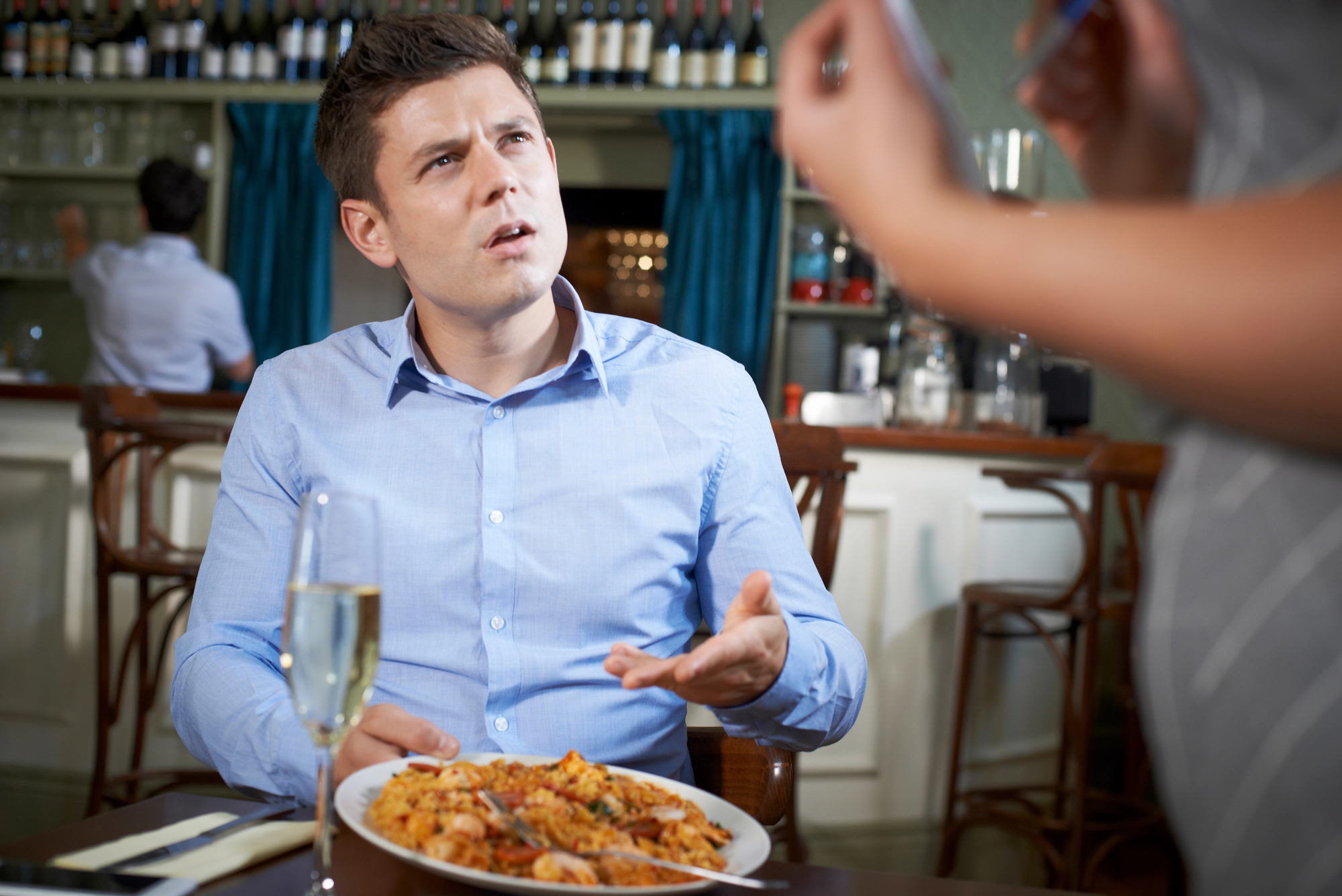 A man in a blue shirt is sitting at a restaurant table with a plate of food, looking confused or frustrated as he talks to a female server who is out of focus. The table has a glass of wine. The background shows shelves with wine bottles and glasses.