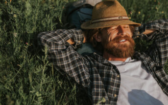 A bearded man wearing a straw hat, plaid shirt, and white t-shirt is lying in tall grass with his arms behind his head and eyes closed, enjoying the sunlight. A backpack is visible beside him.