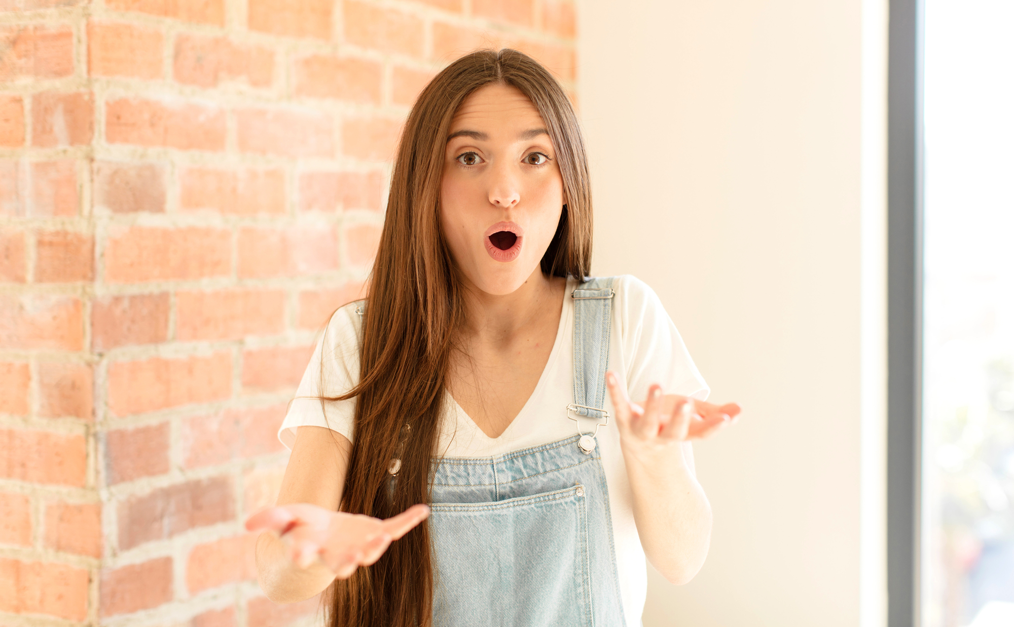 A woman with long brown hair stands in front of a brick wall, wearing a white shirt and denim overalls. She has an expression of surprise on her face, with her mouth open and hands raised.