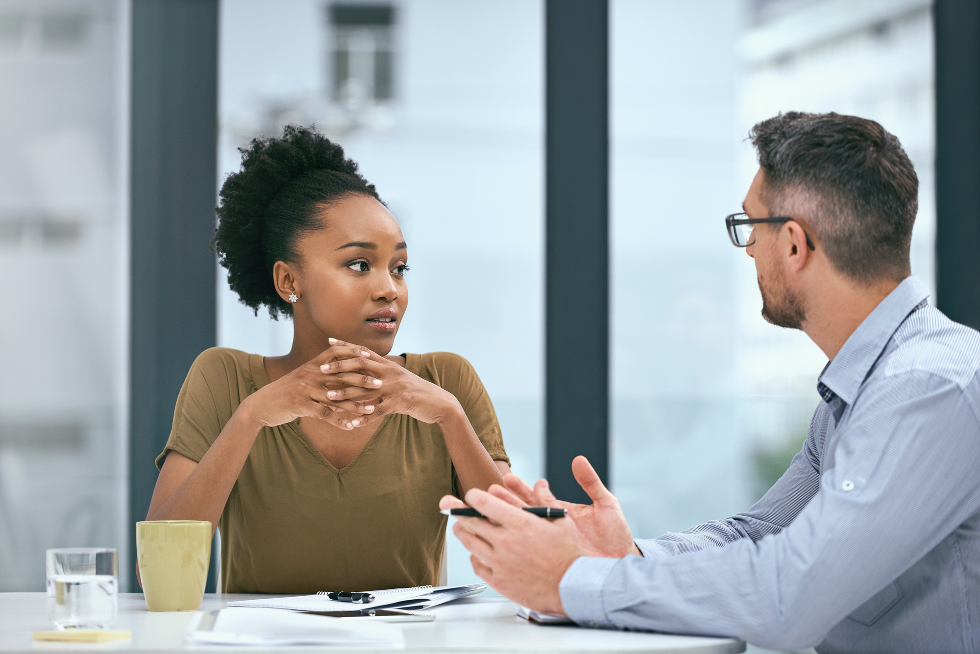 Two people are sitting at a table having a discussion. The woman, with curly hair pulled back, is listening attentively with her hands clasped. The man, wearing glasses and holding a pen, is speaking to her. Papers and a mug are on the table.