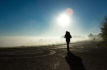 A person walks along a foggy road with trees and light mist in the background under a clear blue sky. The sun shines brightly, casting long shadows on the road. The person is partially silhouetted against the bright light.