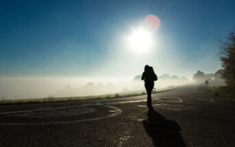 A person walks along a foggy road with trees and light mist in the background under a clear blue sky. The sun shines brightly, casting long shadows on the road. The person is partially silhouetted against the bright light.