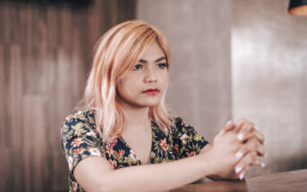 A person with shoulder-length blonde hair and a serious expression is seated at a wooden table. They are wearing a floral patterned shirt and have their hands clasped together, looking ahead thoughtfully. The background features a blurred, warm-toned interior.