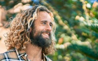 A man with long, curly hair and a beard is smiling while looking to the side outdoors. He is wearing a plaid shirt. The background is filled with greenery, indicating he is in a park or garden on a sunny day.