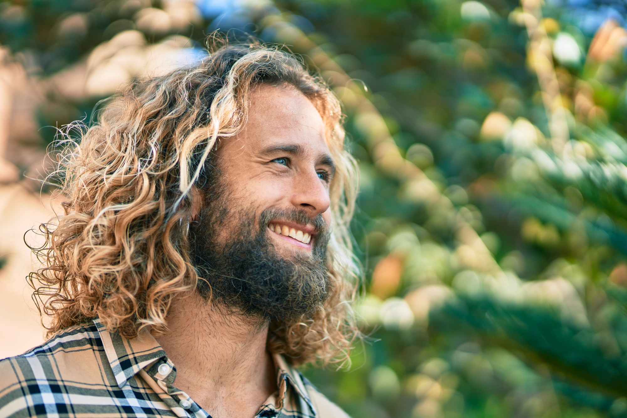 A man with long, curly hair and a beard is smiling while looking to the side outdoors. He is wearing a plaid shirt. The background is filled with greenery, indicating he is in a park or garden on a sunny day.