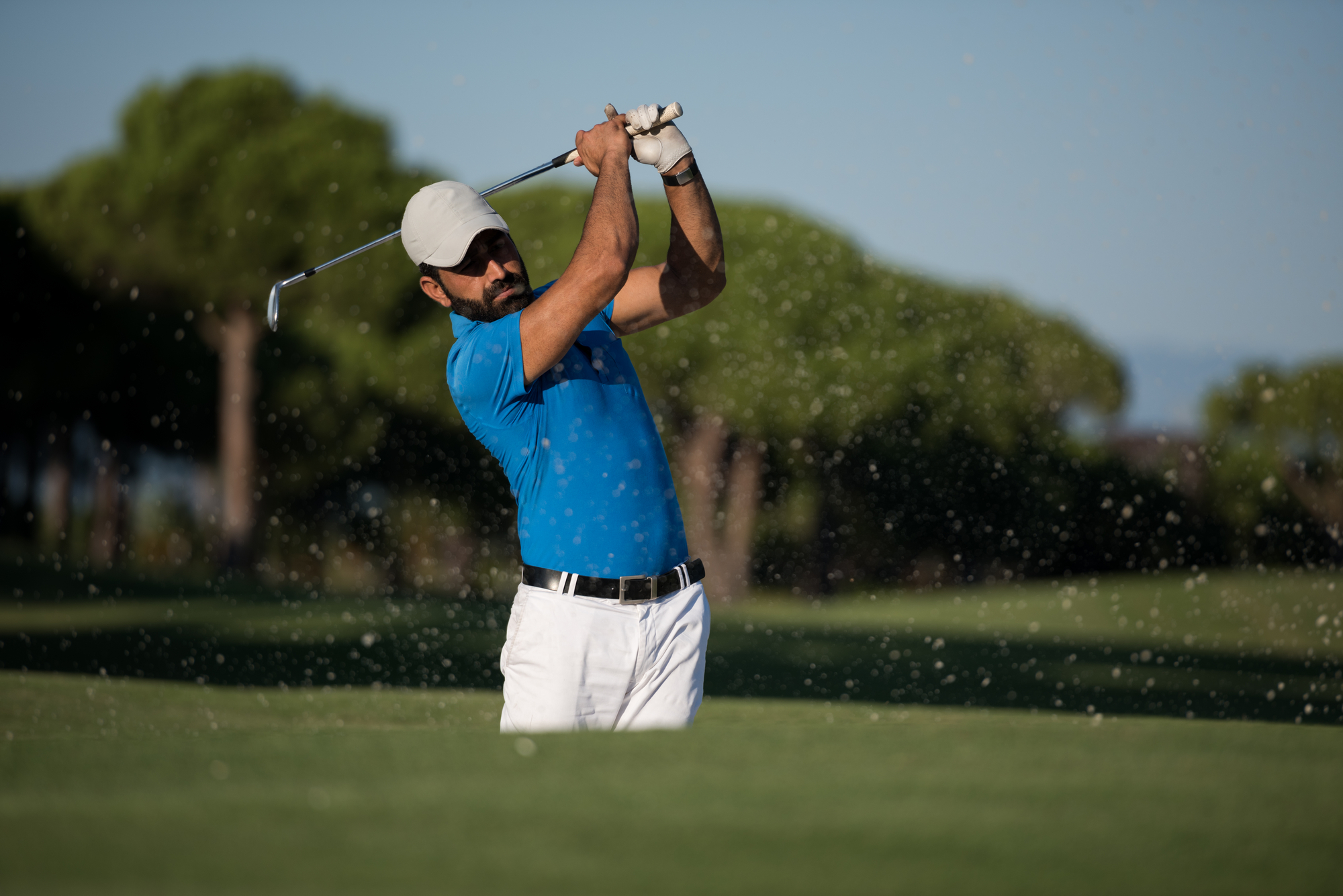 A golfer wearing a white cap, blue polo shirt, and white pants is swinging a golf club in a green, well-maintained golf course. Sand is flying up from the sand trap, suggesting he is hitting the ball out of a bunker. Trees are visible in the background.