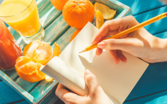 A person's hand holding a pencil and writing in a blank notebook while sitting at a blue table with peeled oranges, a glass of orange juice, and another red drink on a wooden tray. The sunlight casts a warm glow on the scene.