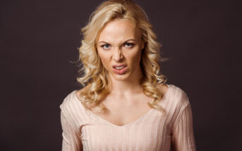 A person with long, wavy, blonde hair and a light pink top is standing against a dark background. They are making an angry, disgusted facial expression with their nose wrinkled and mouth slightly open.