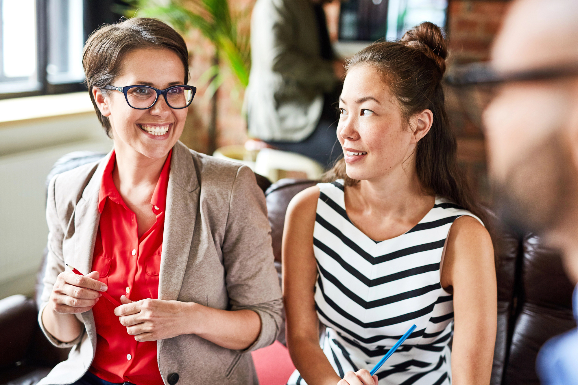 Two women sitting and smiling during a conversation in a well-lit room. One wears glasses, a beige blazer, and a red blouse; the other wears a black and white striped dress. They appear to be engaged and happy. Blurred figure and greenery in the background.