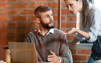 A bearded man sitting at a desk with a laptop looks up at an upset woman gesturing toward him. They are in a modern office with a brick wall background. The man appears surprised or confused, while the woman seems to be expressing frustration.