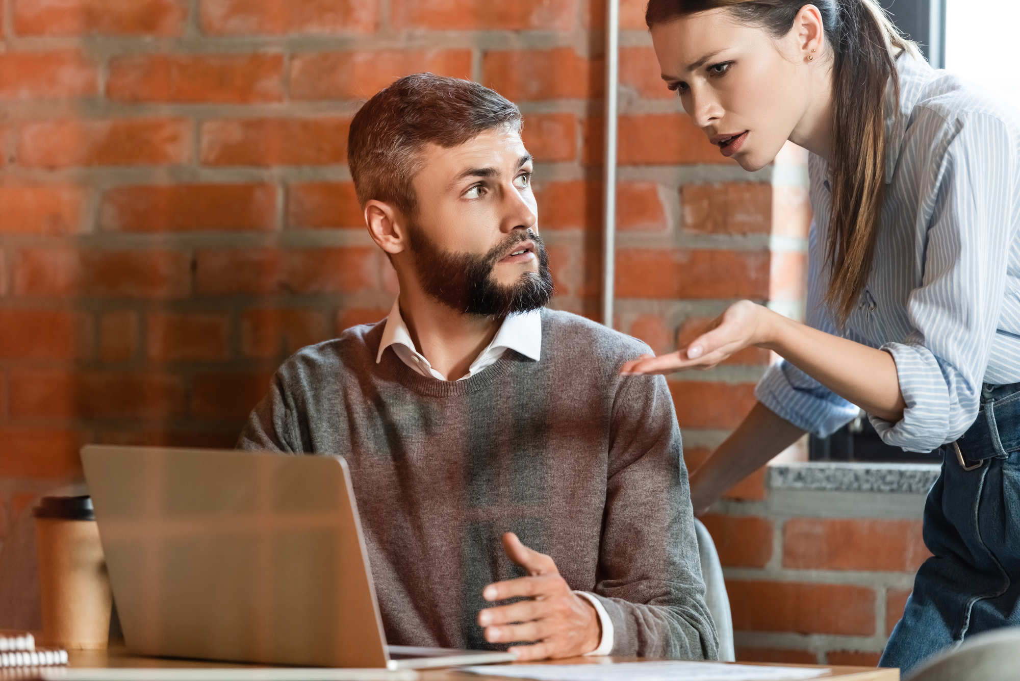 A bearded man sitting at a desk with a laptop looks up at an upset woman gesturing toward him. They are in a modern office with a brick wall background. The man appears surprised or confused, while the woman seems to be expressing frustration.