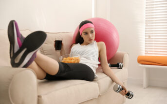 A woman in workout attire lounges on a beige sofa with a pink exercise ball. She holds a dumbbell in one hand and a bowl of snacks in the other, while a drink is placed nearby. She appears to be taking a break from exercising.