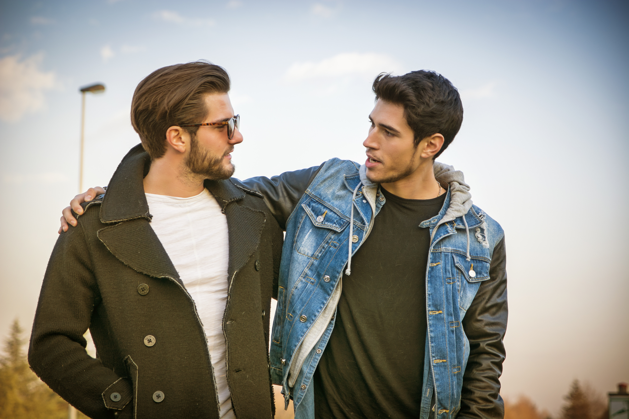 Two young men engage in a friendly conversation outdoors. One wears sunglasses and a dark coat, while the other sports a denim jacket with a black hoodie. Both appear relaxed and amiable under a clear sky.