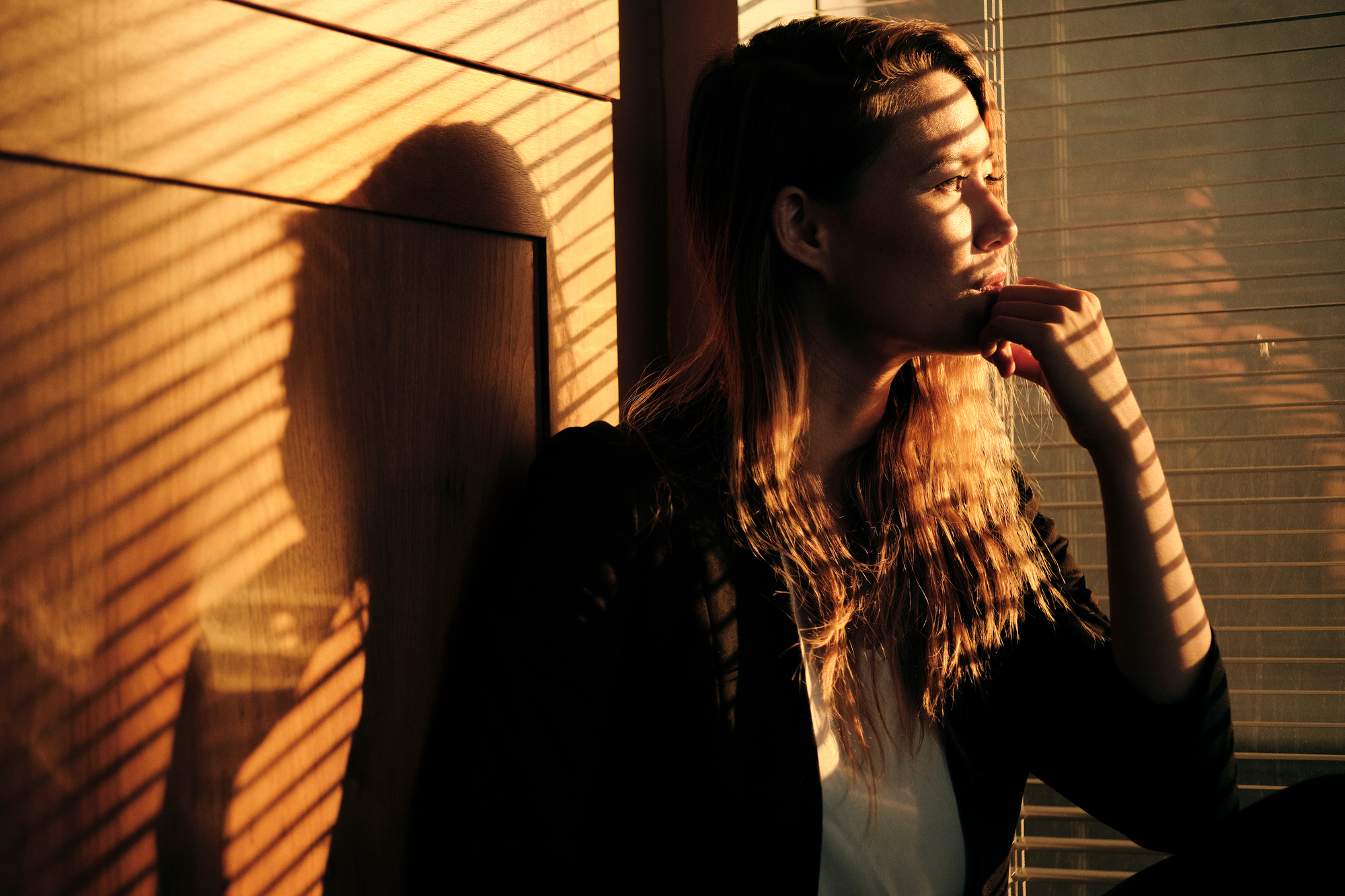 A woman with long hair sits pensively against a wall, bathed in warm sunlight streaming through blinds. The light creates striped shadows across her face and her reflection can be seen in a nearby window.