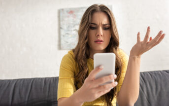 A young woman with long brown hair, wearing a yellow shirt, sits on a grey couch. She holds a smartphone in one hand and looks at the screen with a confused and frustrated expression, her other hand raised in a questioning gesture. A painting is blurred in the background.