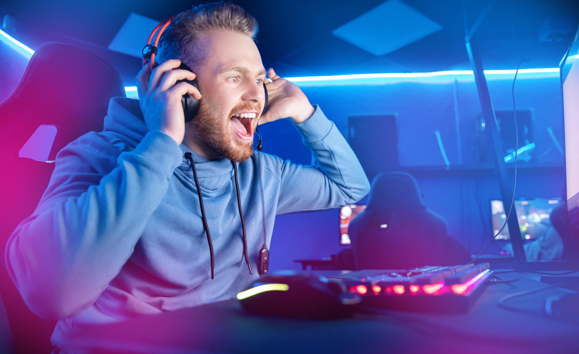 A person wearing a headset and a hoodie sits at a gaming setup with neon lighting. They are enthusiastically looking at a computer screen, one hand adjusting the headset and the other likely on the mouse. The ambiance is energetic with vibrant lighting.