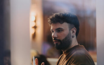 A man with a beard and wavy hair, wearing earrings and a brown shirt, is holding a pen and appears deep in thought. The background is softly blurred, giving a warm ambiance with soft lighting.