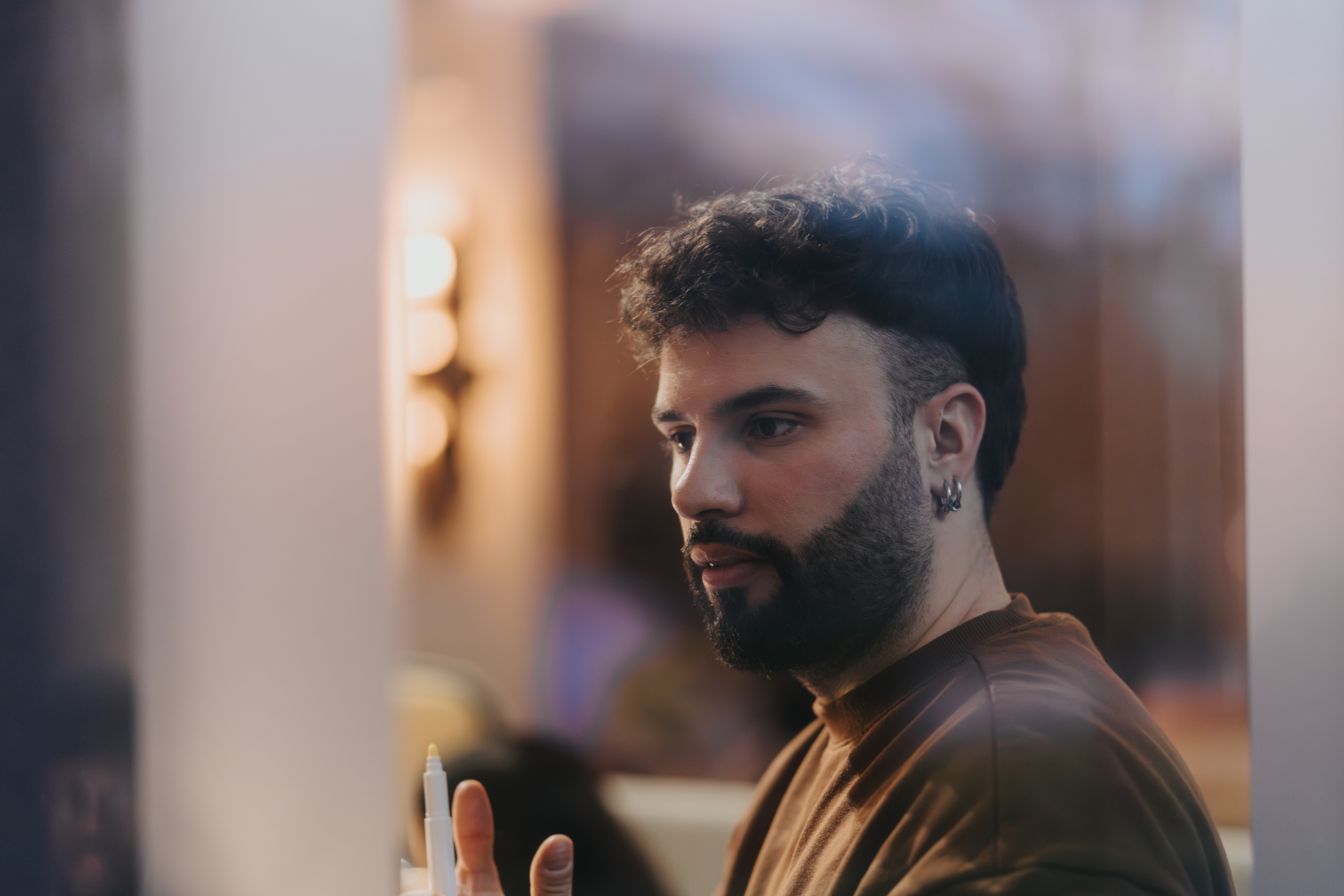 A man with a beard and wavy hair, wearing earrings and a brown shirt, is holding a pen and appears deep in thought. The background is softly blurred, giving a warm ambiance with soft lighting.