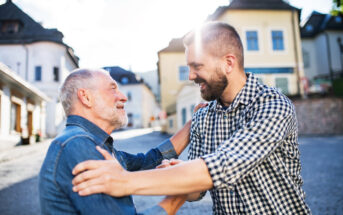 Two men, one older and one younger, face each other with broad smiles in an outdoor setting. The older man has gray hair and a beard, wearing a denim shirt. The younger man has a beard and is wearing a checkered shirt. They are greeting each other warmly, with hands on each other's shoulders. Bright sunlight and charming buildings can be seen in the background.