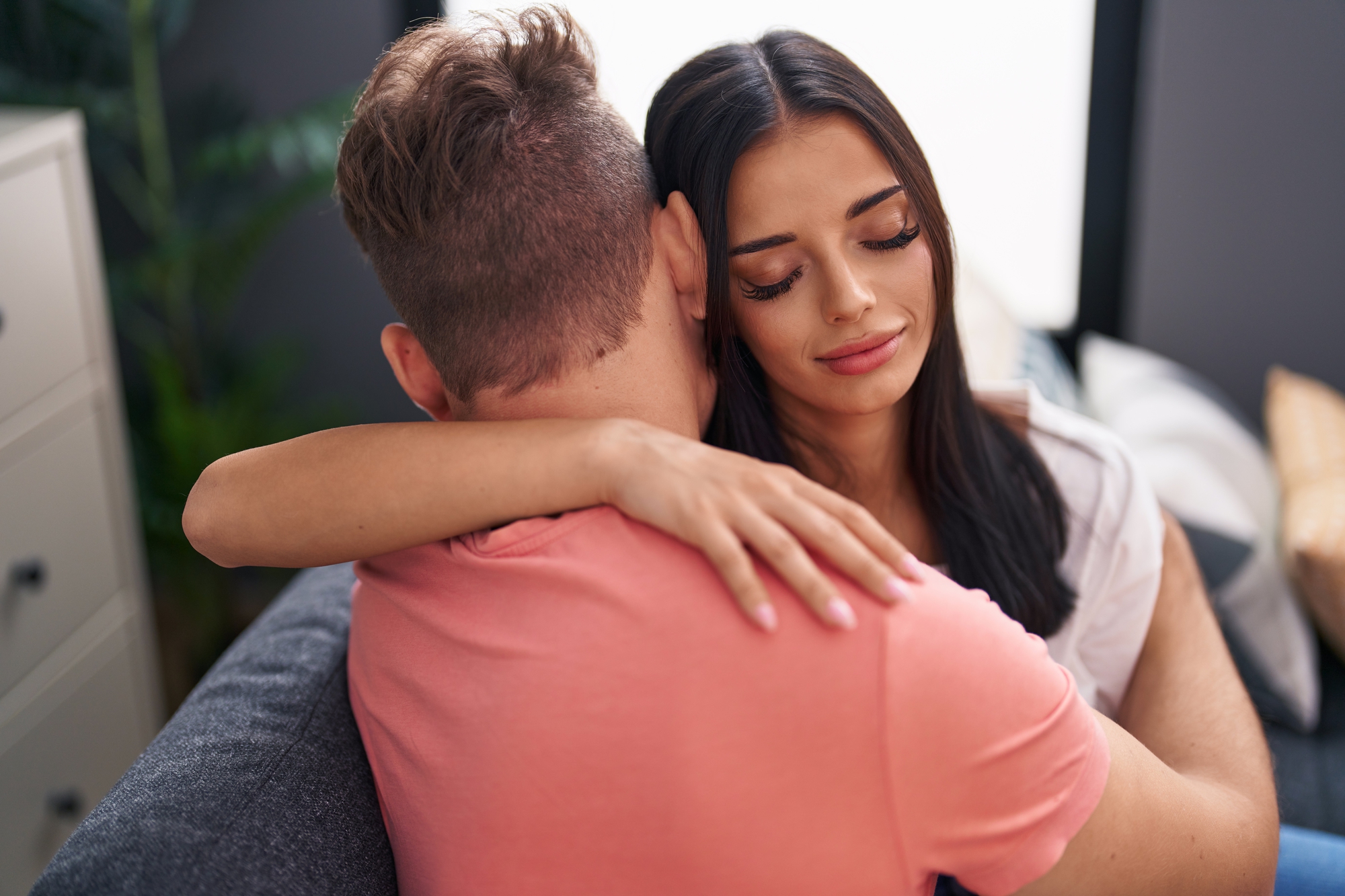 A woman with long dark hair embraces a man wearing a pink shirt while they sit on a couch. The woman has a serene expression with her eyes closed, resting her head on the man's shoulder, creating an intimate and comforting moment.