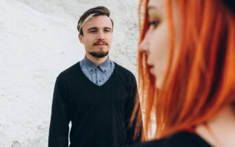 A man with short hair and a beard, wearing a black sweater over a blue patterned button-up shirt, is looking forward with a neutral expression. In the foreground, a woman with vibrant red hair, slightly out of focus, is partially visible in profile.