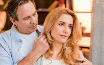 A middle-aged man affectionately adjusts the hair of a middle-aged woman with a thoughtful expression. They stand close together in a warmly lit room, creating an intimate, serene atmosphere.