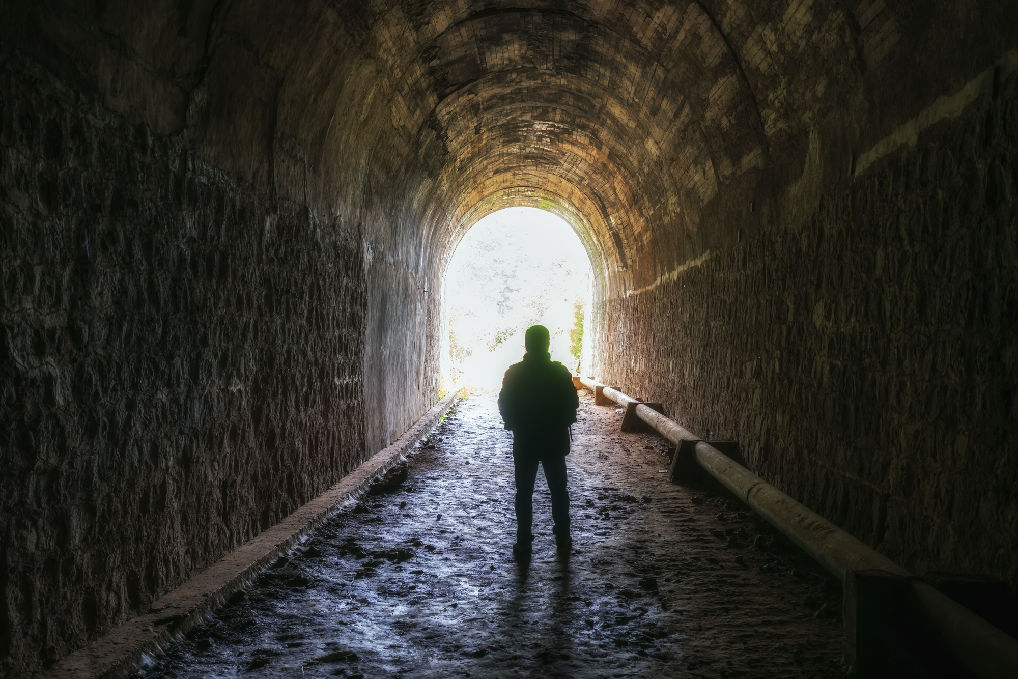 A silhouette of a person standing alone in the middle of a dimly lit tunnel. The tunnel walls are rugged, and the ground is uneven. Bright light is visible at the tunnel's end, indicating the exit. The scene evokes a sense of mystery and adventure.